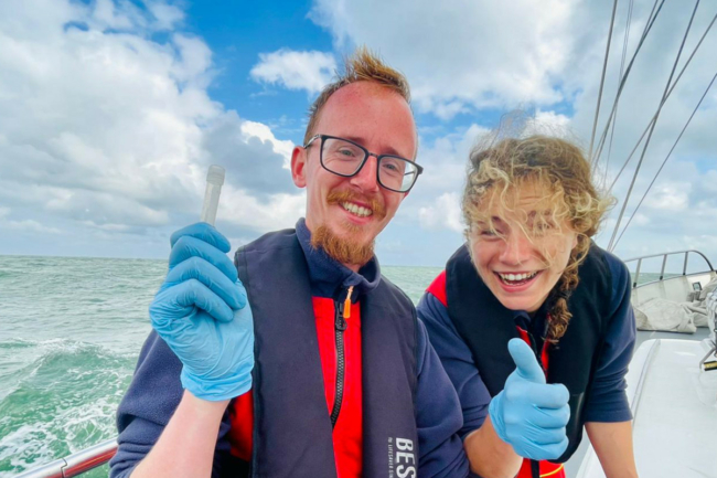 two Sea Rangers, one male, one female, pose on a ship. The male is holding up a test tube, and the female is giving a thumbs up to the camera.