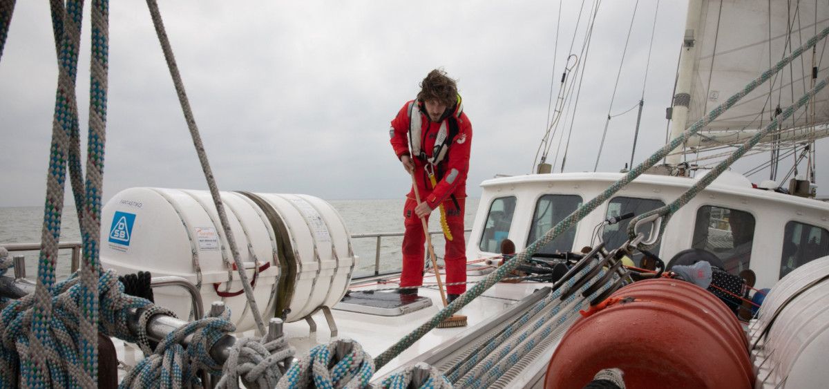 A lone Sea Ranger in uniform is on the deck of a ship, cleaning it with a long-handled brush. The sky and sea in the background are gray.