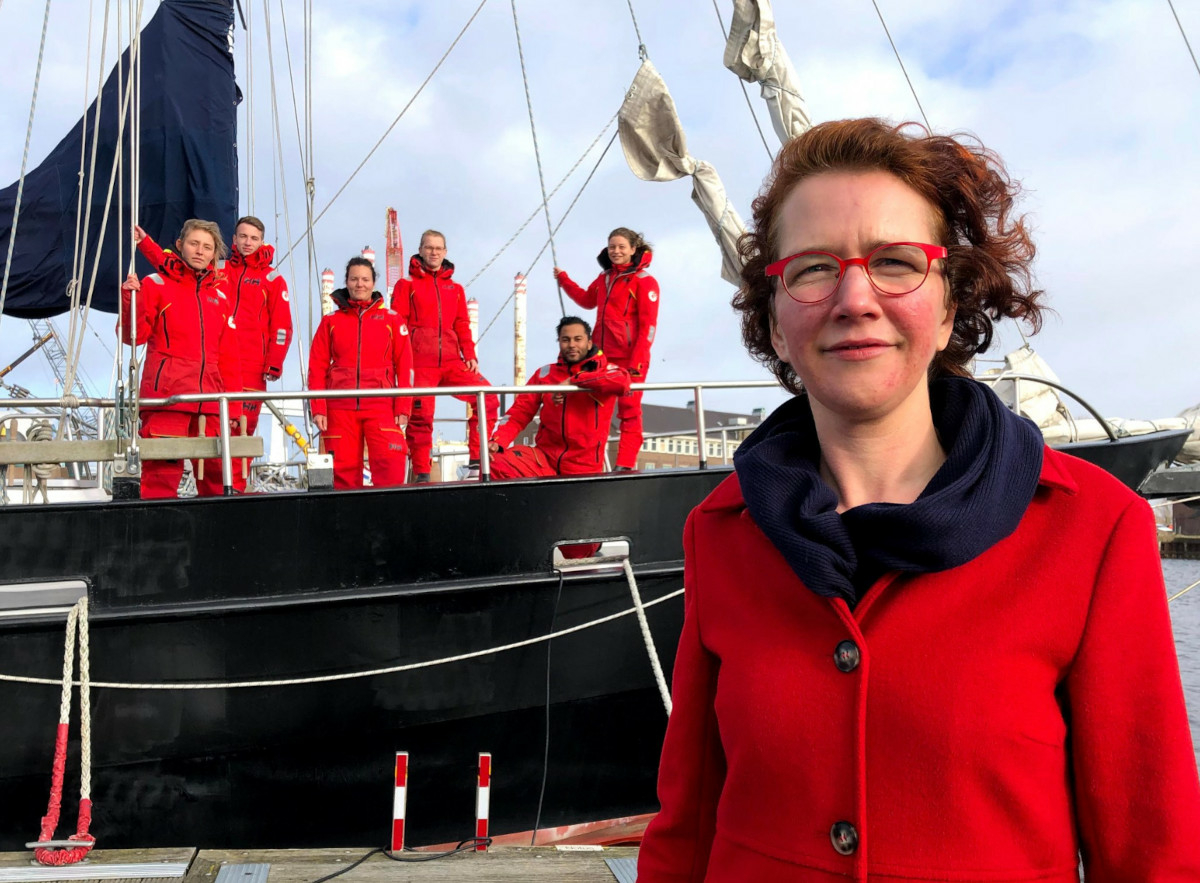 The new managing director of Sea Rangers Service, Christel Pullens, stands in a red coat in front of a black ship. On the ship, six Sea Rangers stand in uniform. It is a sunny cloudy day.