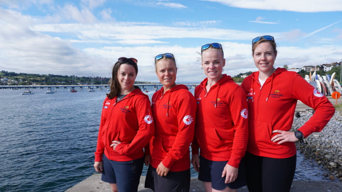 Four female Sea Rangers in red tops and blue shorts smile and pose in front of a body of water. Small sailing boats can be seen on the water.