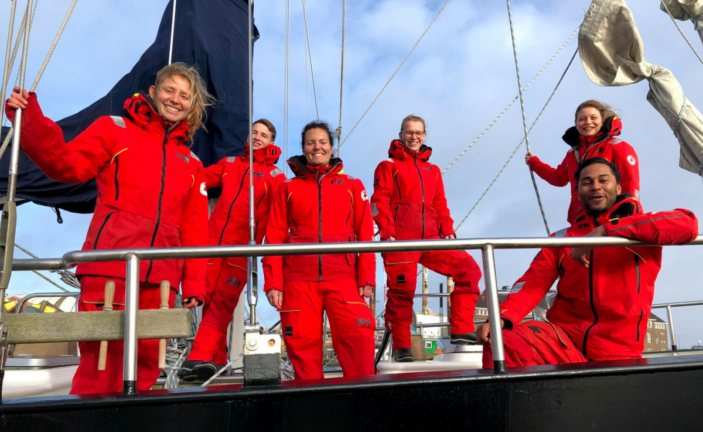 Six Sea Rangers in red uniform are onboard a Sea Ranger ship. All are looking towards the camera, smiling. In the background, you can see some buildings and the blue sky.