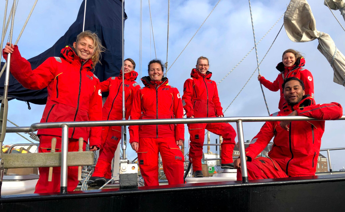 Six Sea Rangers in red uniform are onboard a Sea Ranger ship. All are looking towards the camera, smiling. In the background, you can see some buildings and the blue sky.
