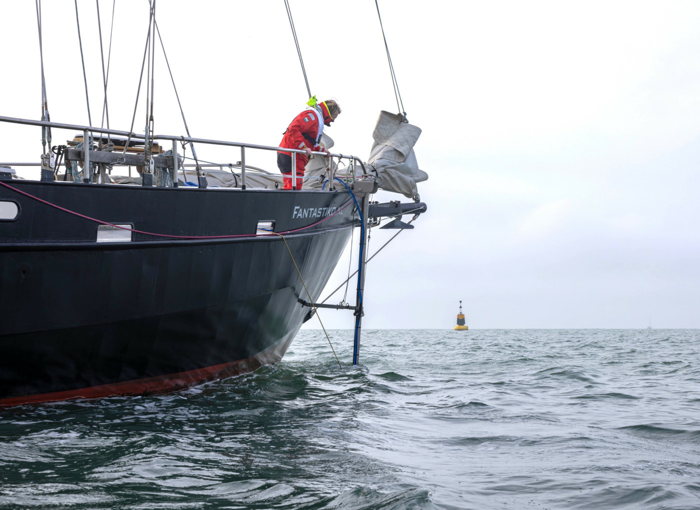 A Sea Ranger dressed in a red uniform is looking over the side of a navy blue ship at the ocean, undertaking hydrographic surveying.
