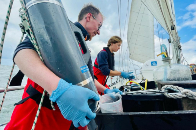 Two Sea Rangers on board a ship stood in front of scientific equipment measuring pollution levels in water samples as part of their ocean conservation work.